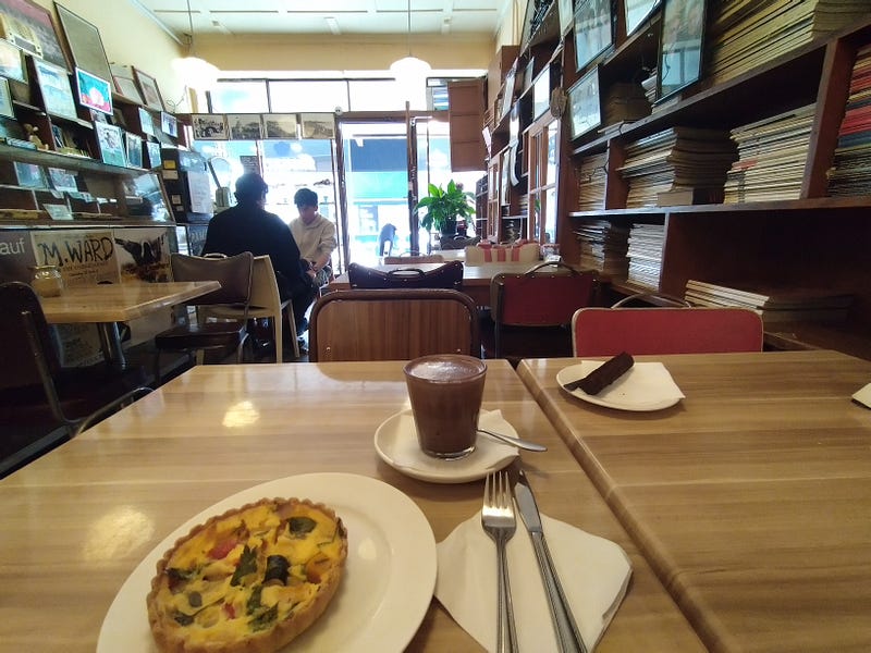 A wide photo of a table in a narrow wooden café, where bookshelves and posters cover the walls. On the table, a quiche, hot chocolate and chocolate-coated marzipan loaf wait to be devoured.