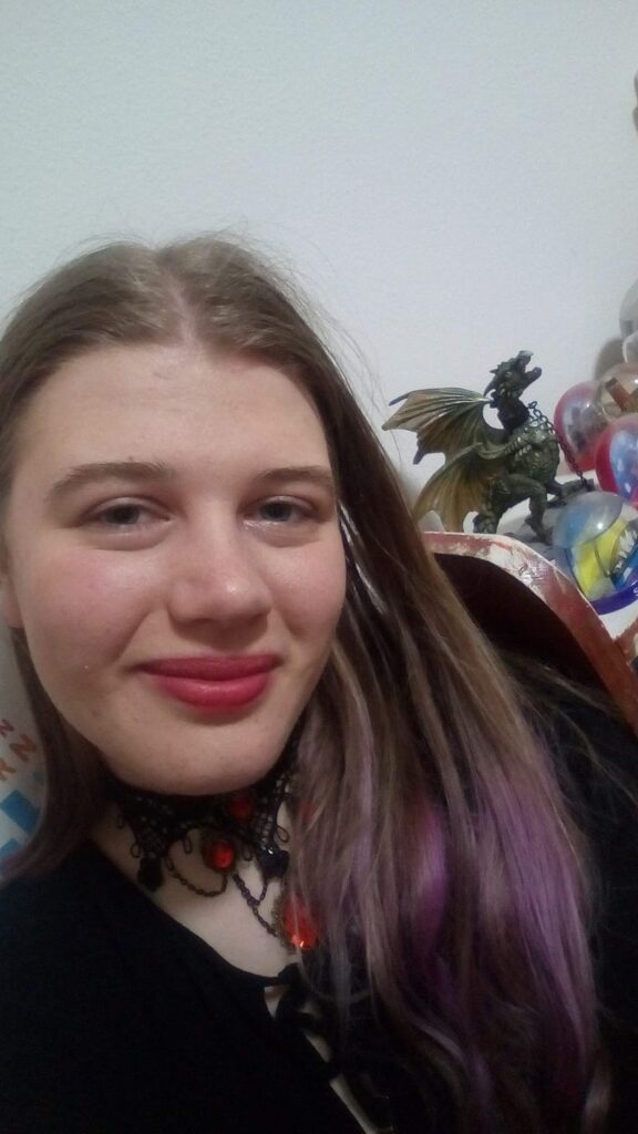 Selfie of a femme teen with purple at the end of their dirty blonde wearing a delicate black shirt, a black lace choker with red plastic jewels and a smile in pinkish-red lipstick. They are posing before a shelf with chipping paint, upon which stands a wyvern figurine and at least four snowglobes.