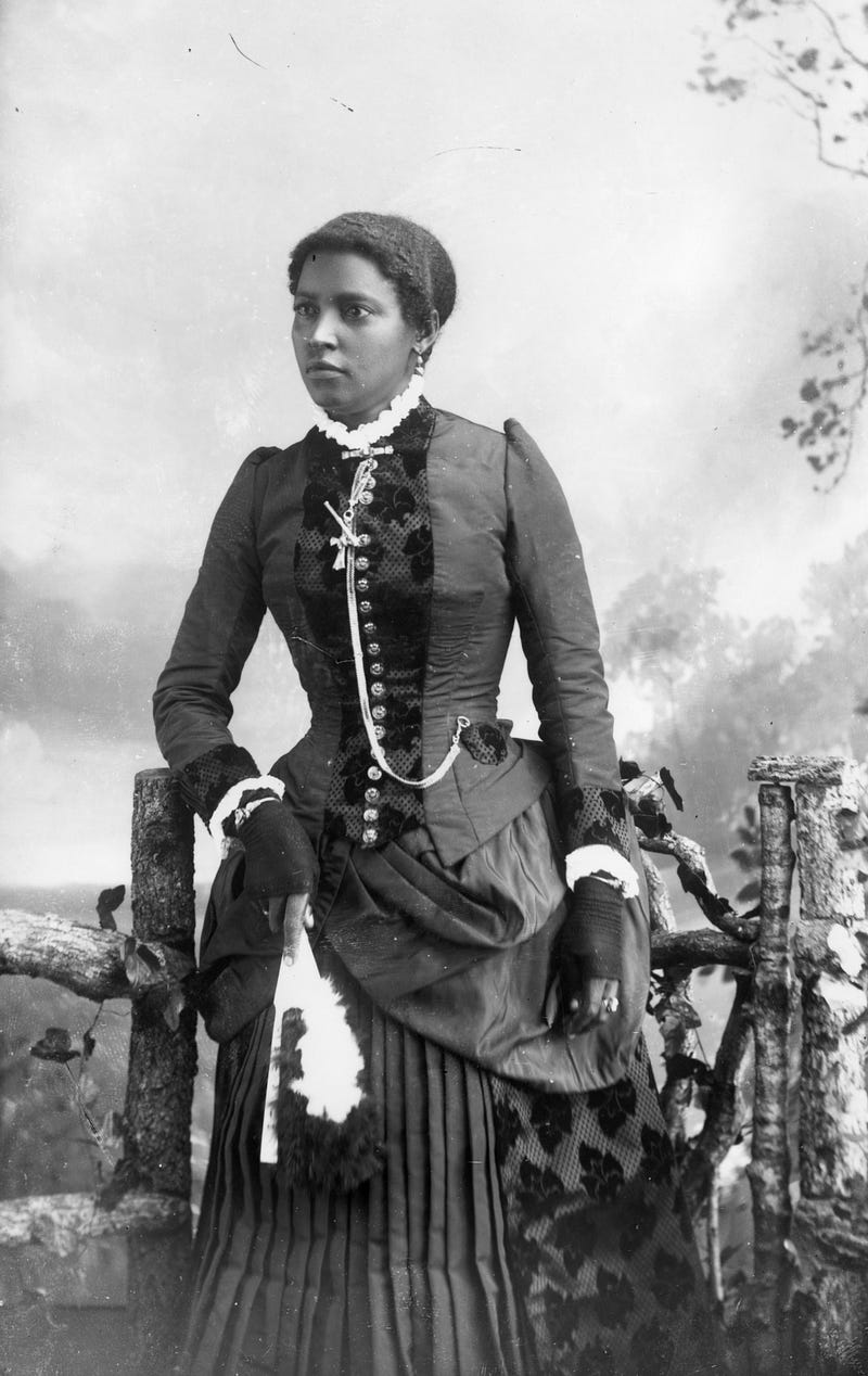 A monochrome photograph of a well-dressed African American woman holding a fan with feathers before a fencepost and a clouded sky.