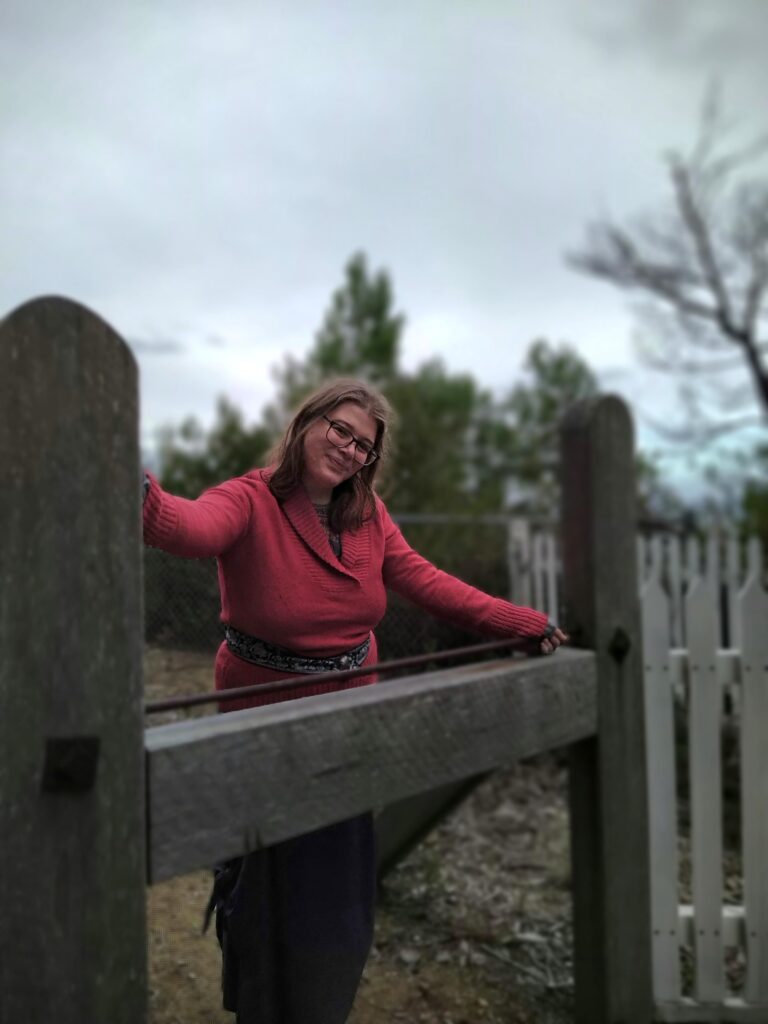 Sarah Rosina Winkler, age 25: a curvy femme in a woollen red jumper and grey skirt behind a wooden post before a wind-swept sky.